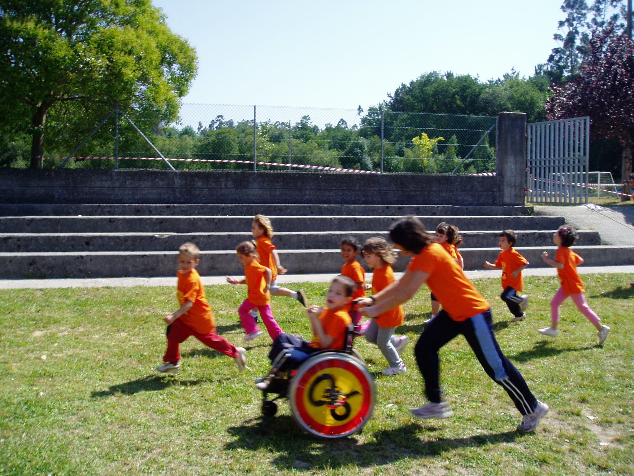Un grupo de niños de infantil participando en una carrera. Todos llevan las mismas camisetas de color naranja chillón. Uno de ellos participa en su silla de ruedas y es la profesora quien la empuja. Todos se sitúan en torno a la silla. Porque el objetivo no es llegar el primero, sino llegar todos juntos.